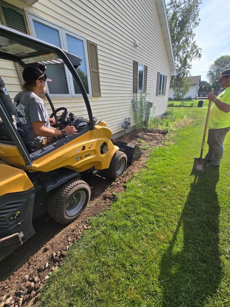 Worker operating a mini excavator for landscaping alongside a colleague with a shovel.