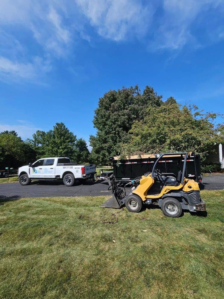 Yellow landscaping vehicle parked on a lawn with a truck in the background under a blue sky.