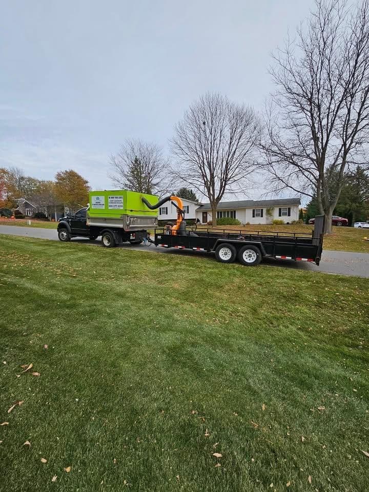 Green truck with a trailer parked on a residential street with trees and homes in the background.