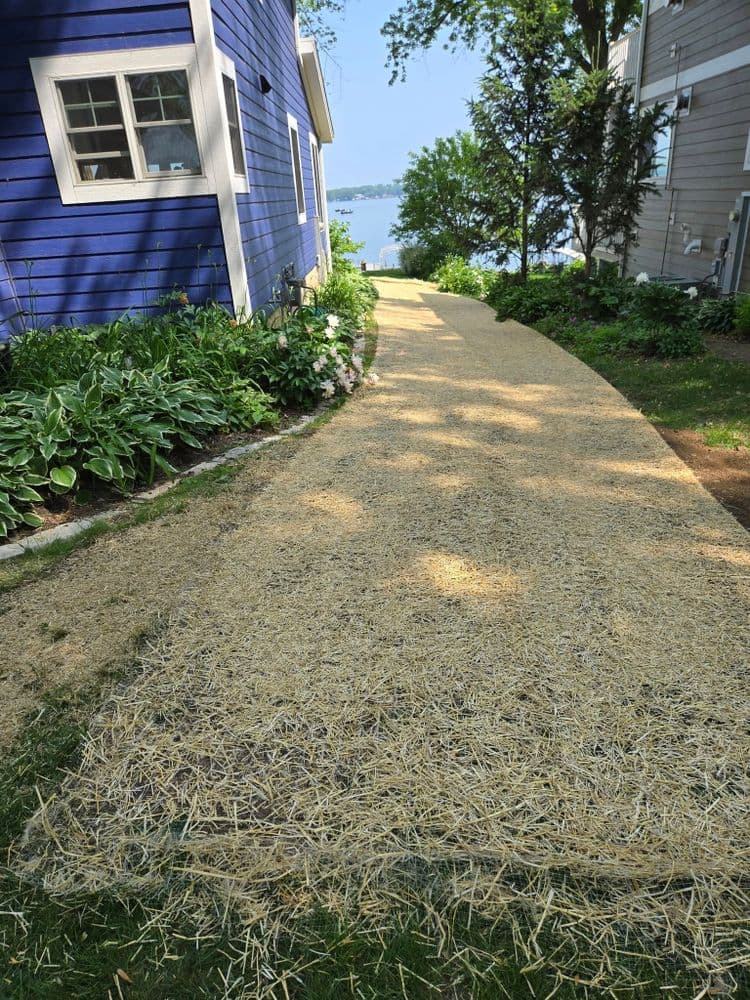 Pathway lined with straw leads to a lake, surrounded by lush greenery and blue house.